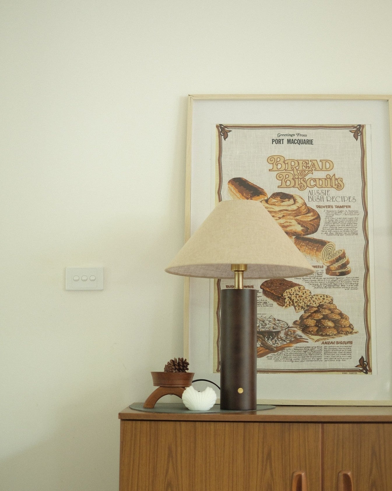 Morgan Table Lamp in Walnut with linen shade on a wooden cabinet with a framed poster of bread in the background.