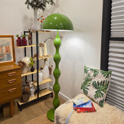 Living room with a Dori Wavy Mushroom Floor Lamp in Olive Green with sculptural wavy base, plants, and decorative items on a shelf.