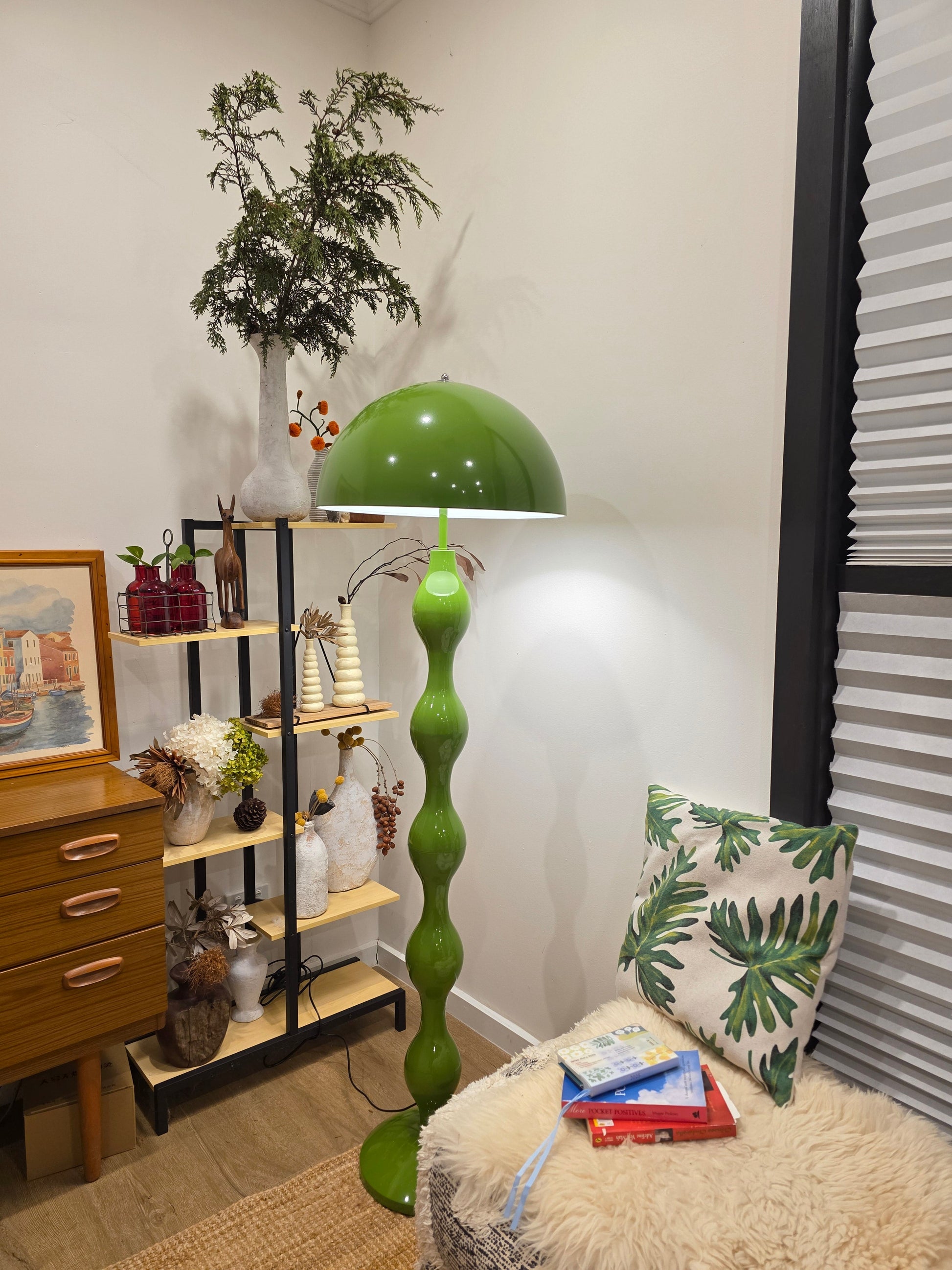Living room with a Dori Wavy Mushroom Floor Lamp in Olive Green with sculptural wavy base, plants, and decorative items on a shelf.