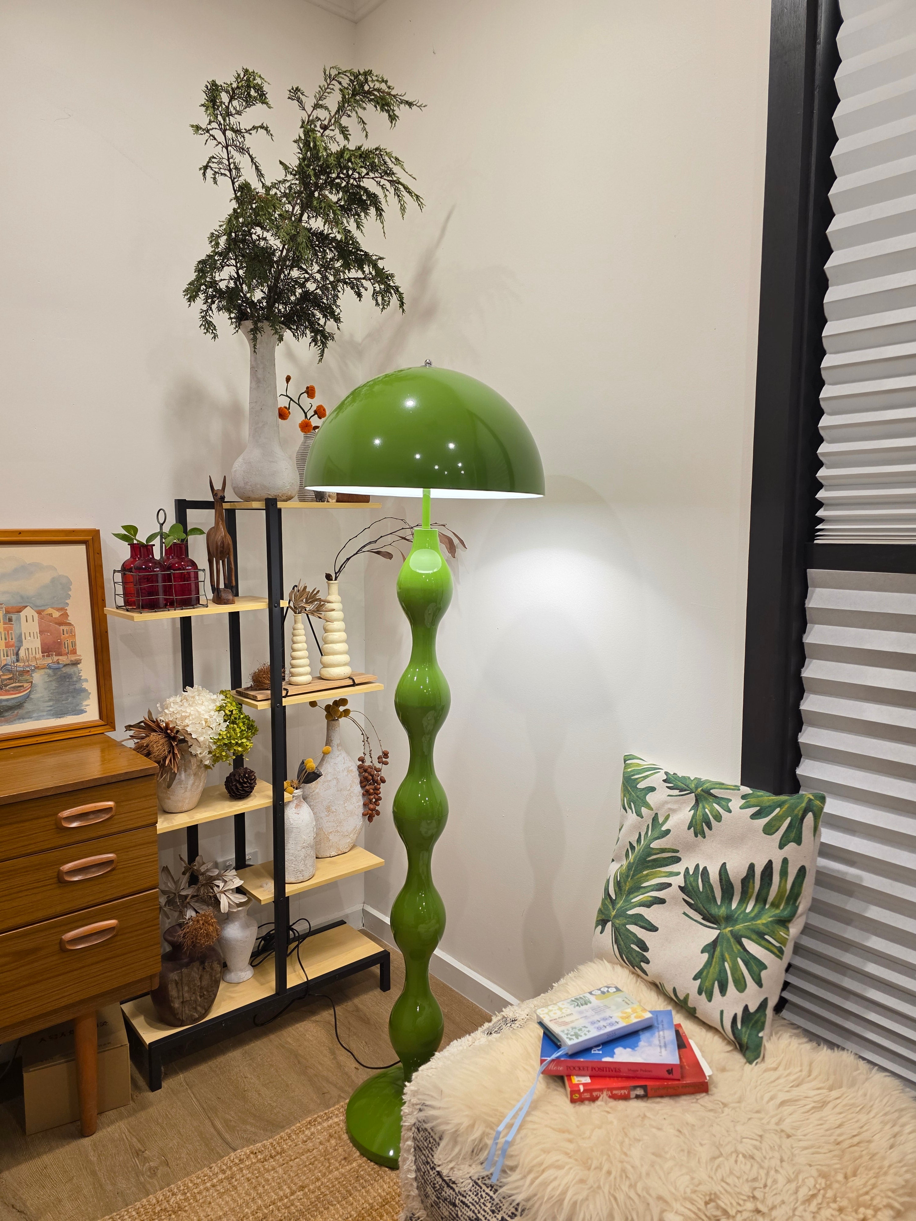 Living room with a Dori Wavy Mushroom Floor Lamp in Olive Green with sculptural wavy base, plants, and decorative items on a shelf.