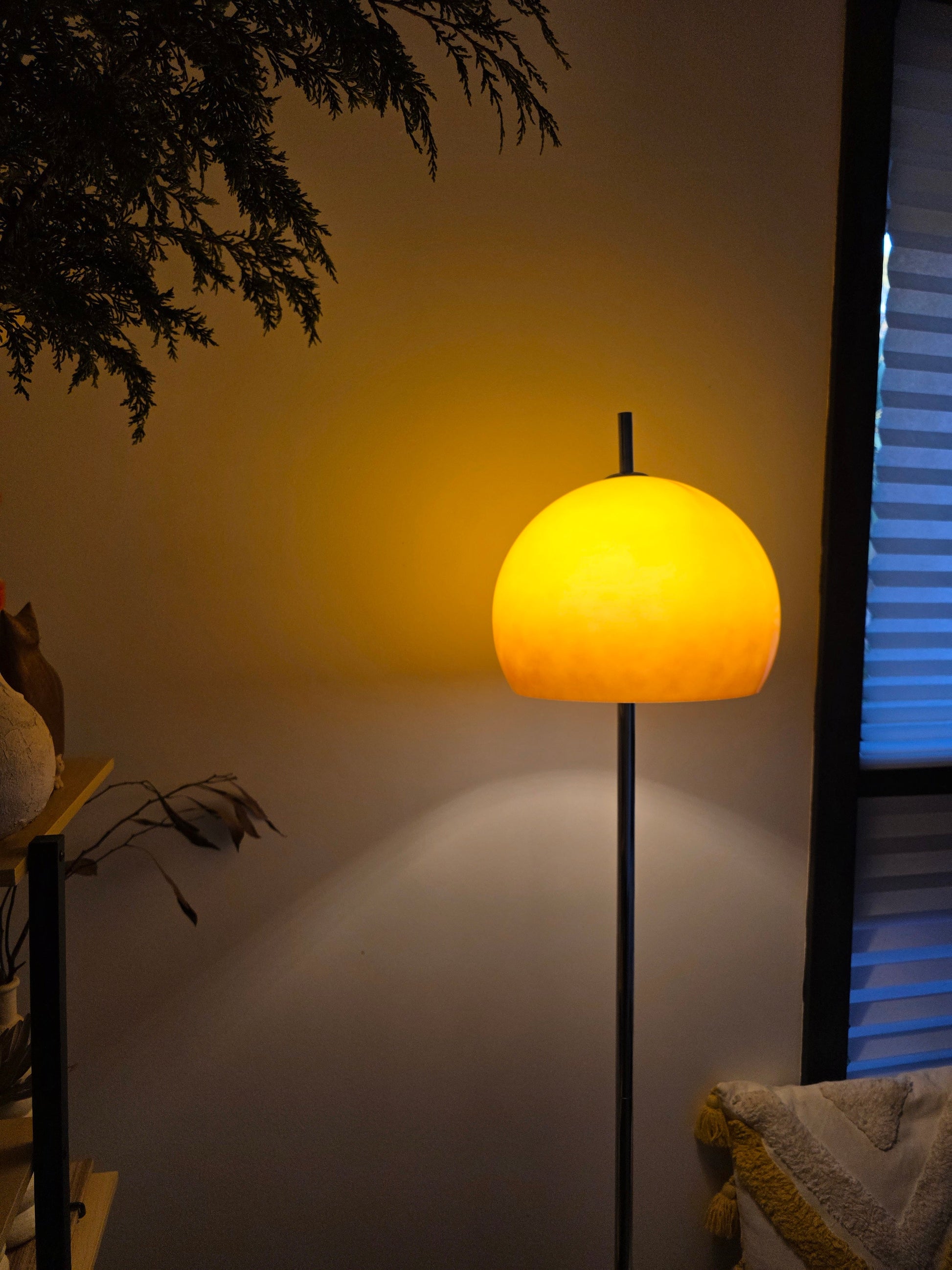 Olivia Mushroom Bauhaus Yellow floor lamp casting a shadow on a wall with plants and a window in the background.