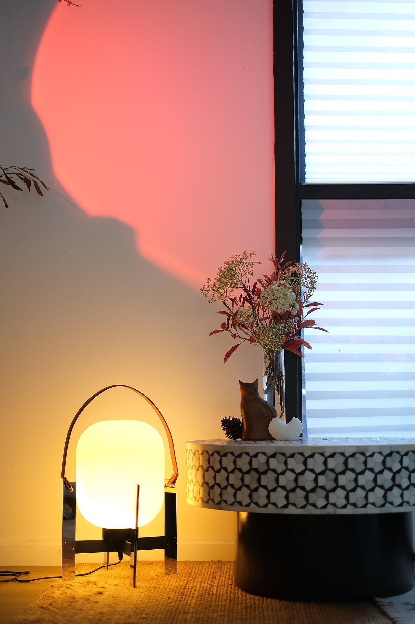 Alix Cesta Metalica Basket Lamp with chrome frame and opal glass globe with a warm glow next to a table with a vase of flowers and a cat, against a window with blinds.