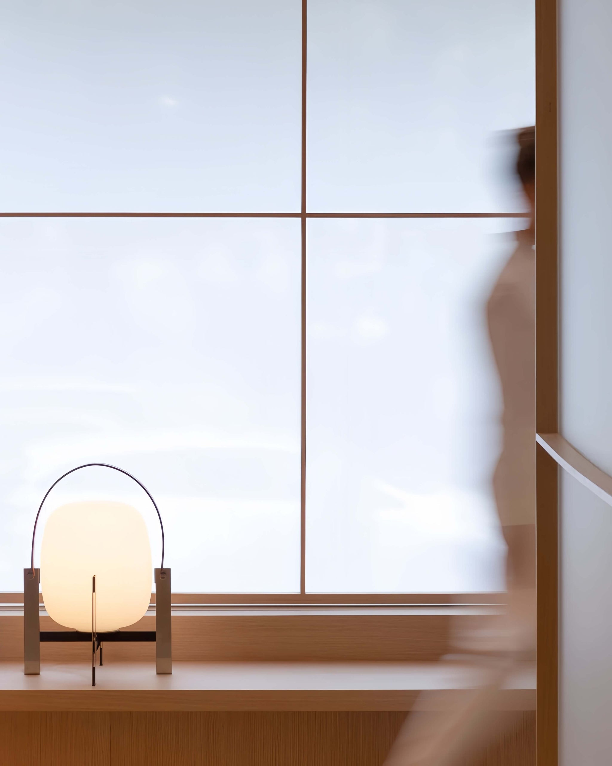 Alix Cesta Metálica Basket Lamp with chrome legs and frosted glass on a wooden surface with a blurred person and window in the background
