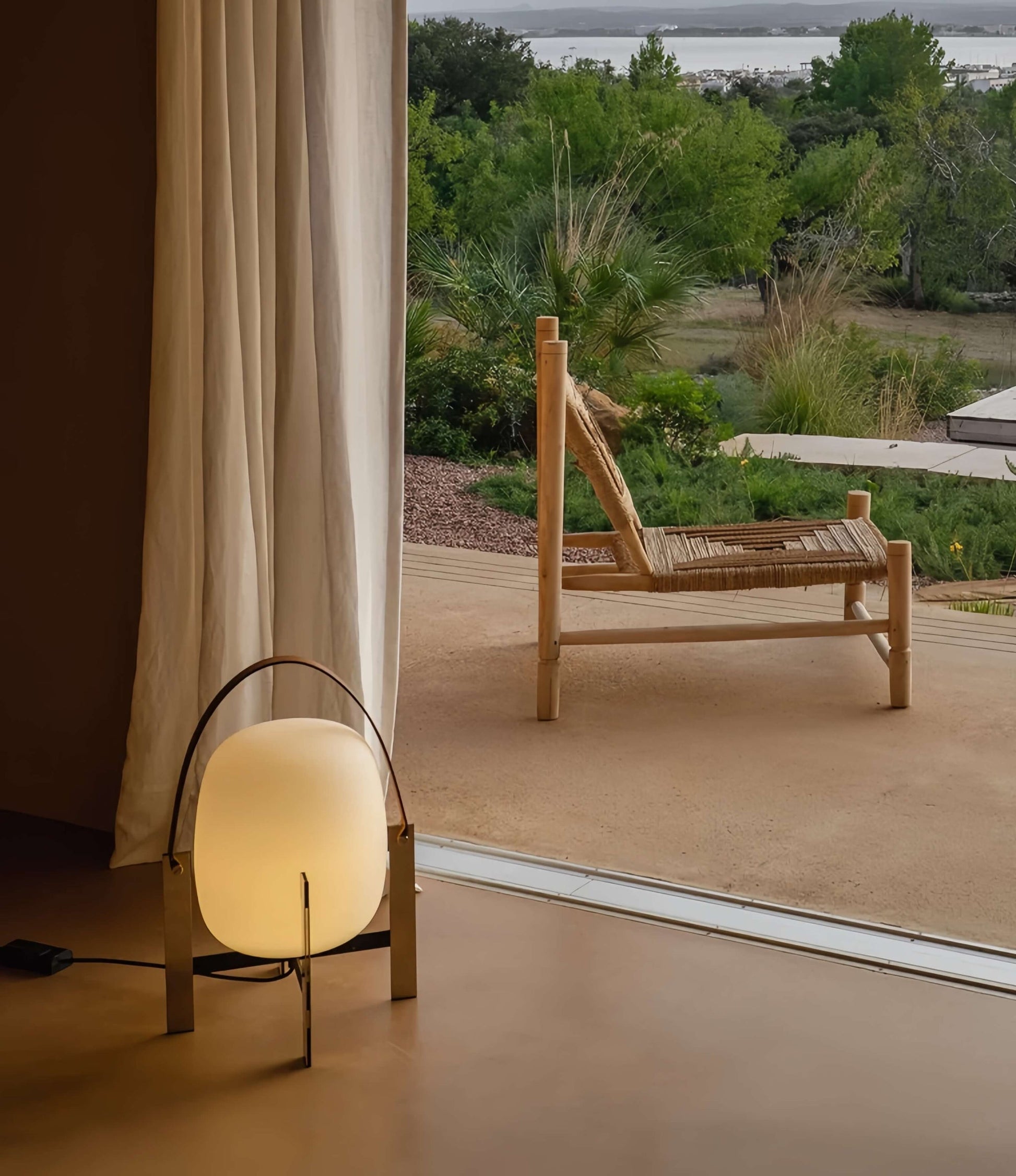 Alix Cesta Metálica Basket Lamp with chrome legs and frosted glass on a floor with a view of a patio and garden through a window.