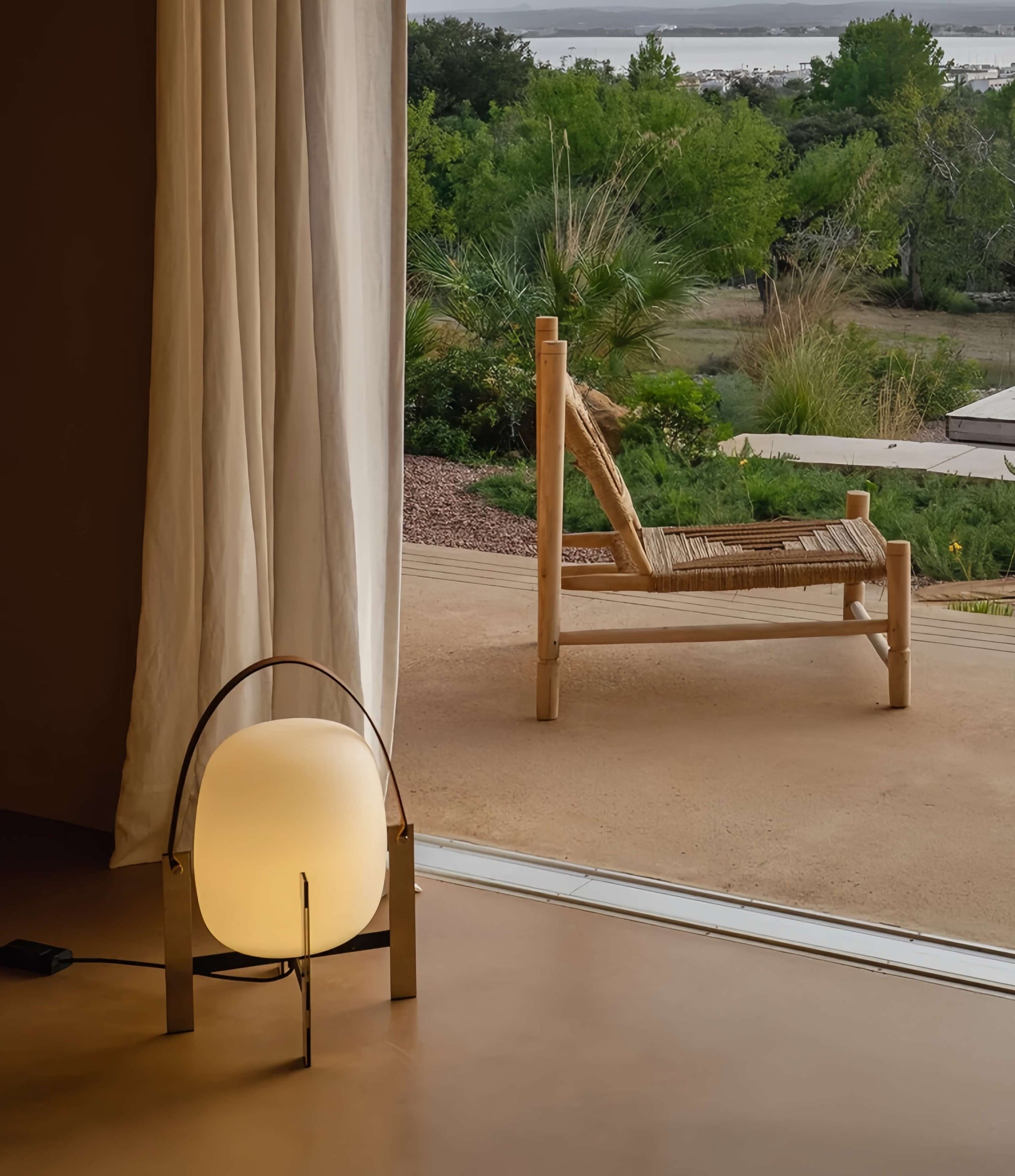 Alix Cesta Metálica Basket Lamp with chrome legs and frosted glass on a floor with a view of a patio and garden through a window.
