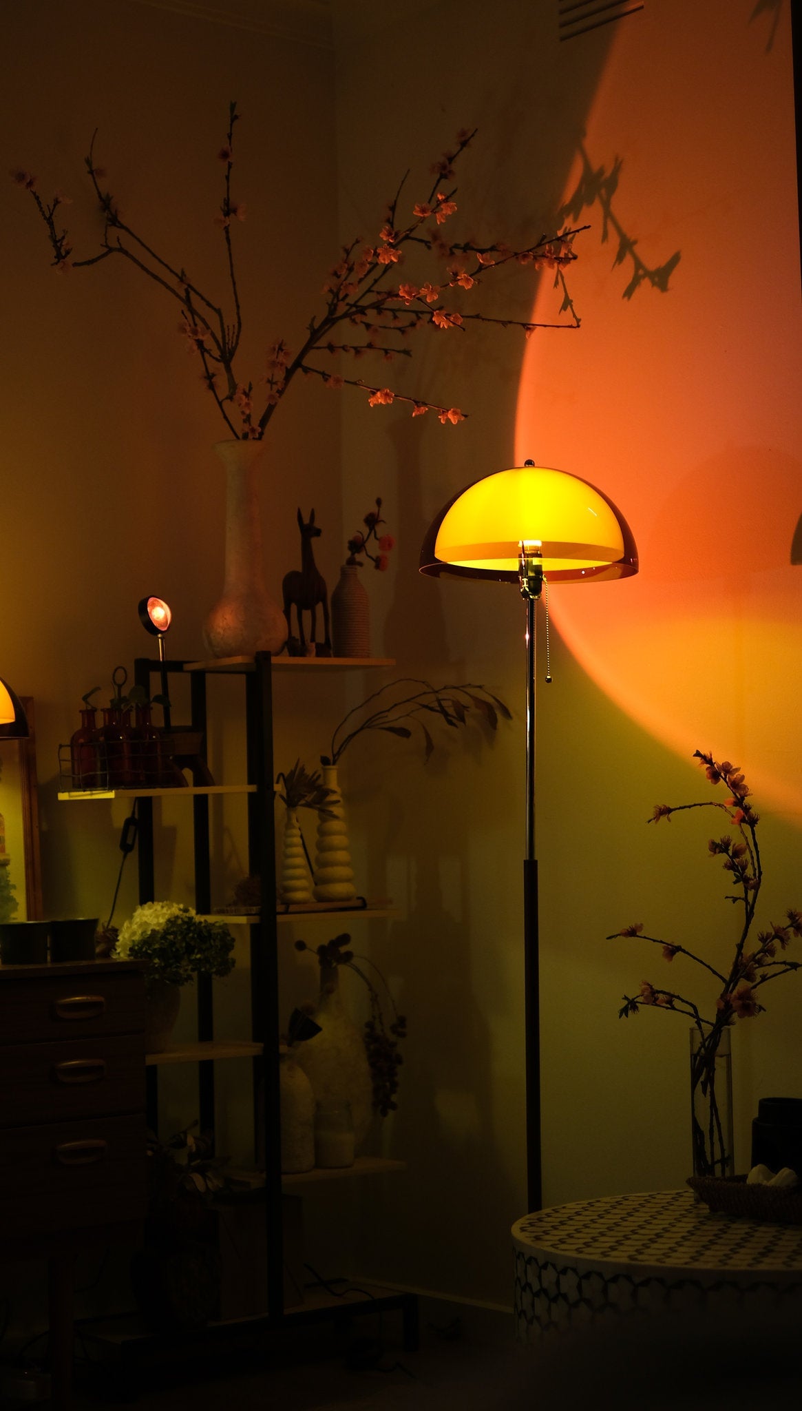 Coby+ Mushroom Bauhaus Floor Lamp Orange with Pull Chain Dark room with a floor lamp casting a warm glow, shelves with decorative items, and plants.