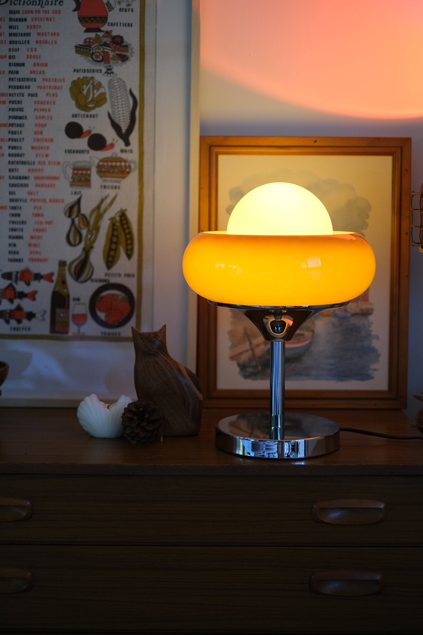 Egg Tart Table Lamp with orange dome glass shade and white inner globe with a glowing orange shade on a wooden surface, with a cat and decorative items in the background.