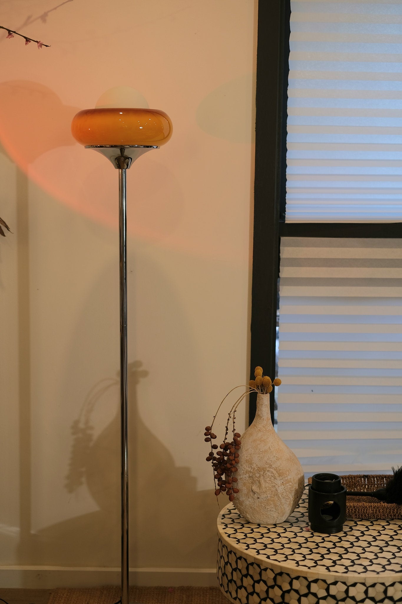 70s inspired Egg Tart lamp styled as cozy corner lighting with a round amber shade next to a small table with a vase and other items against a wall.