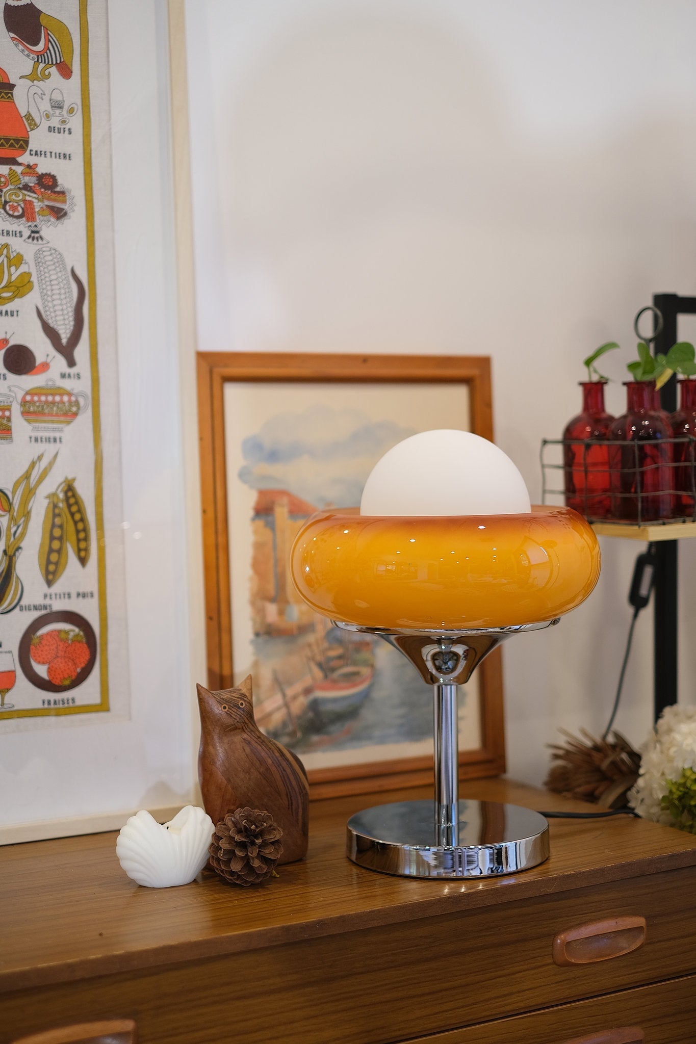 Vintage-style Egg Tart Table Lamp with orange dome glass shade and white inner globe on a wooden surface with decorative items and framed artwork in the background.