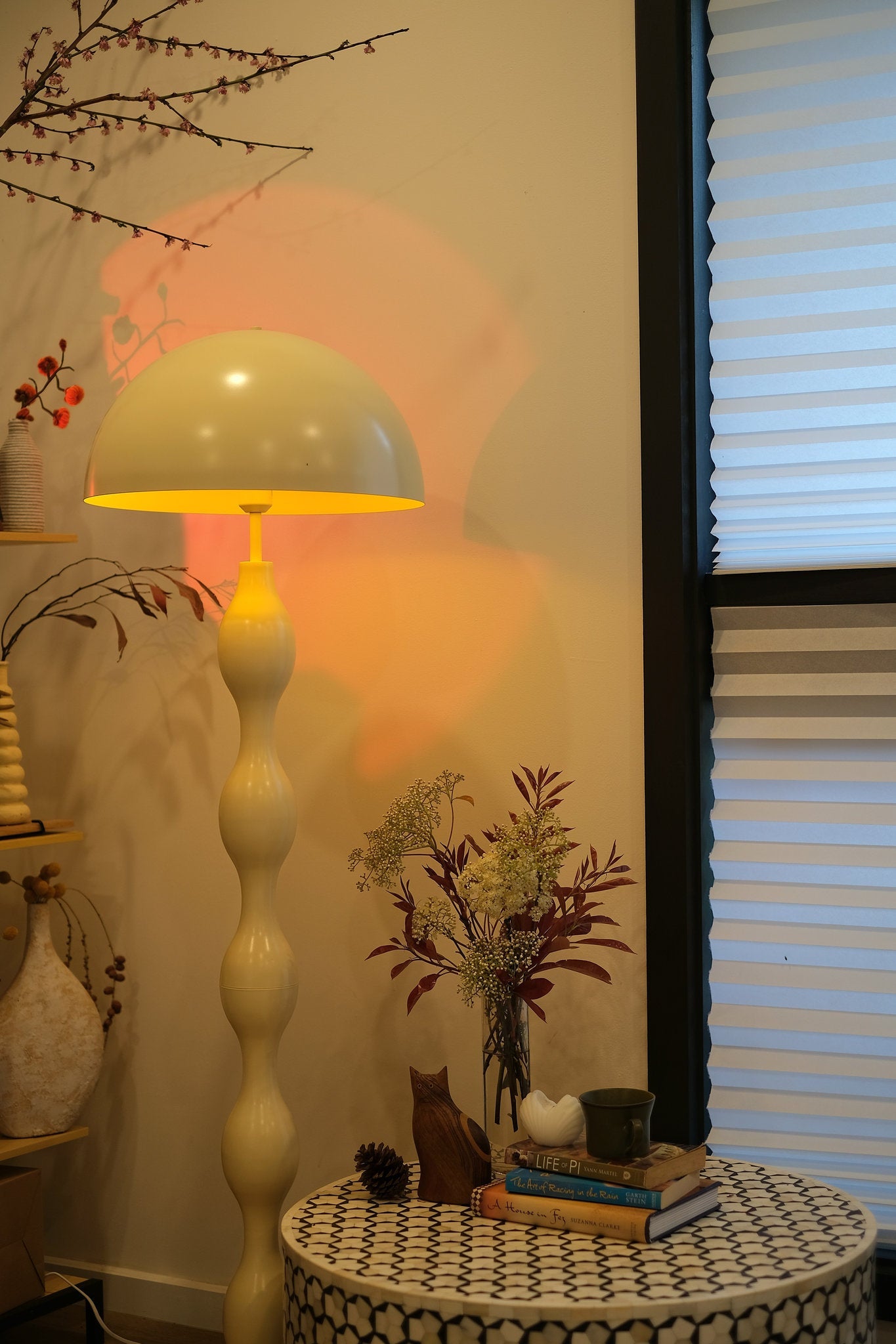 Retro modern mushroom lamp with wavy sculptural base in butter yellow on a patterned table against a wall with a window.