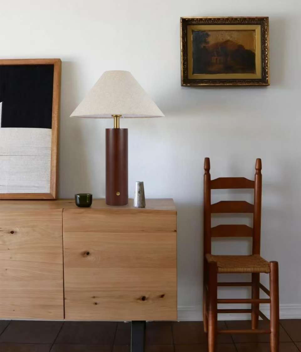 Wooden dresser with a Morgan Table Lamp in Walnut with linen shade on sideboard and chair against a white wall with a painting.