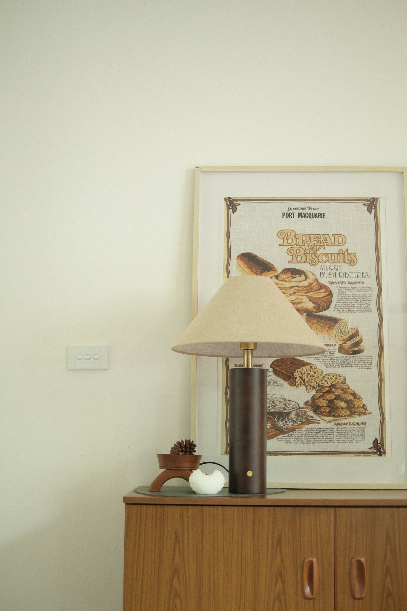 Morgan Table Lamp in Walnut with linen shade on a wooden cabinet with a framed poster of bread in the background.